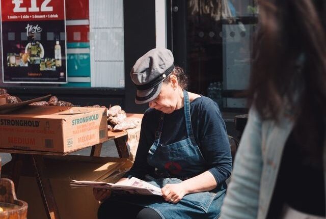 A female vendor reads a newspaper at a bustling outdoor market under a bright red awning.